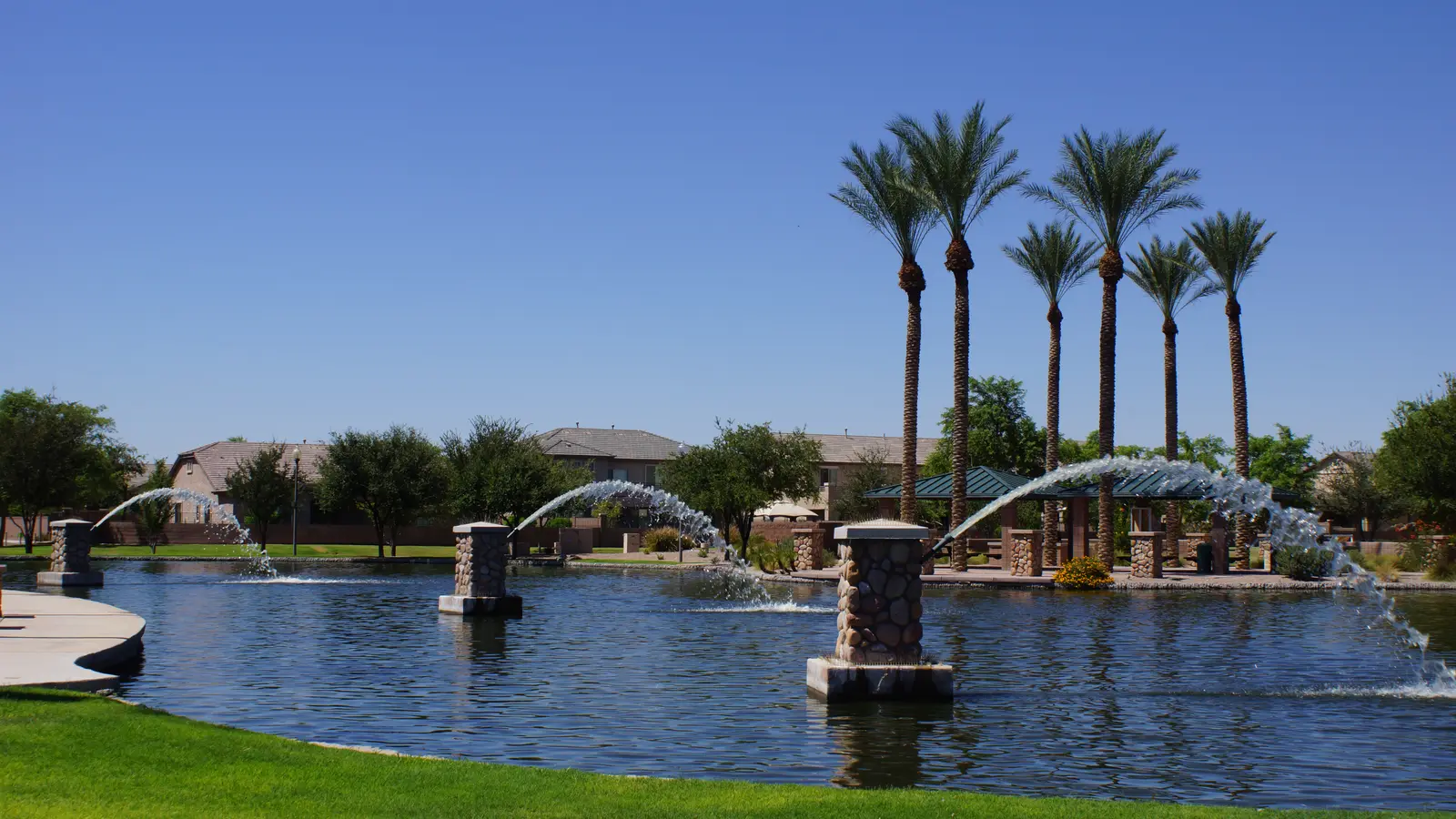 Palm-lined park lake with arcing fountains and tile-roofed homes beyond in Maricopa, Arizona
