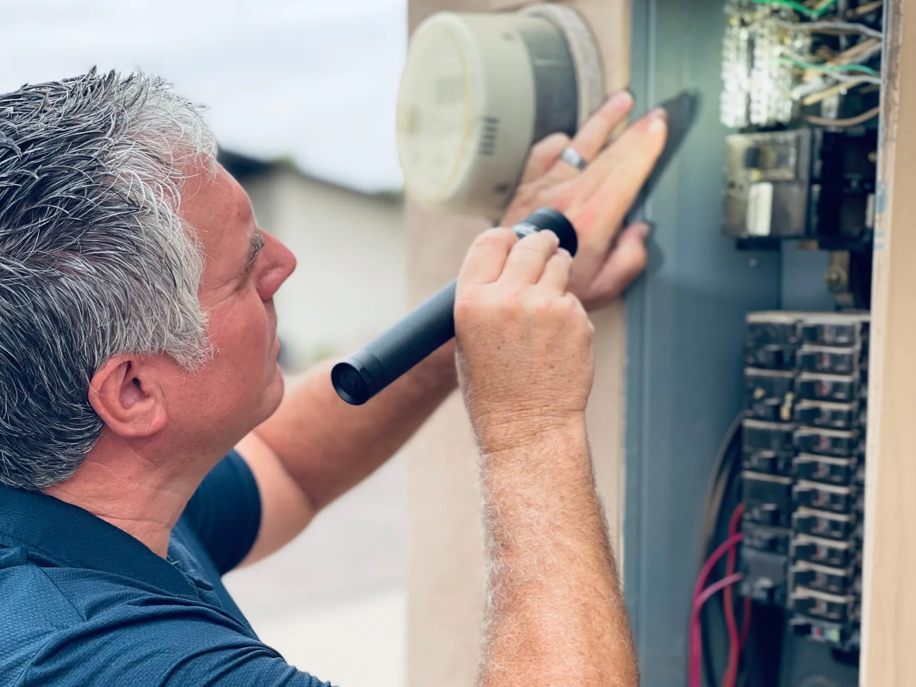 Bob Clardy inspecting an open exterior electrical service panel with a flashlight